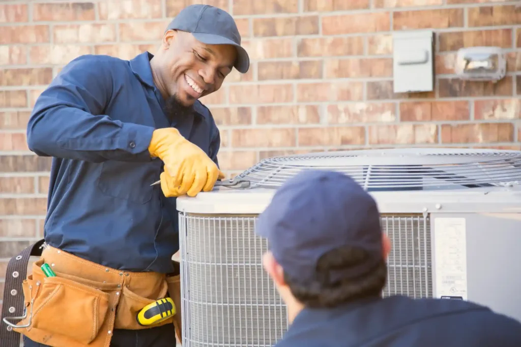 AC Service In Ellenwood, GA 1 HVAC technicians in blue uniforms servicing an outdoor air conditioning unit near a brick wall with electrical meter and conduit