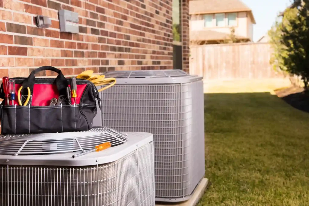 AC Tune-Up In Ellenwood, GA 1 Tool bag with HVAC tools placed on an outdoor air conditioning unit beside a brick house, with a second unit and residential backyard in view